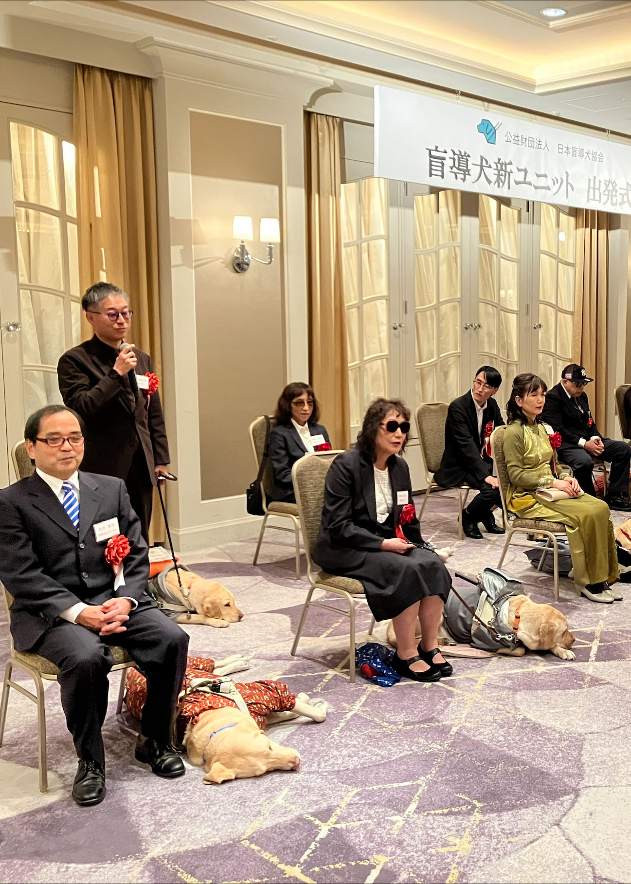 盲導犬ユーザーの出発式のう様子。等間隔に椅子が置かれ、ユーザーと盲導犬が並んでいる。盲導犬は皆ゆったりと伏せている。