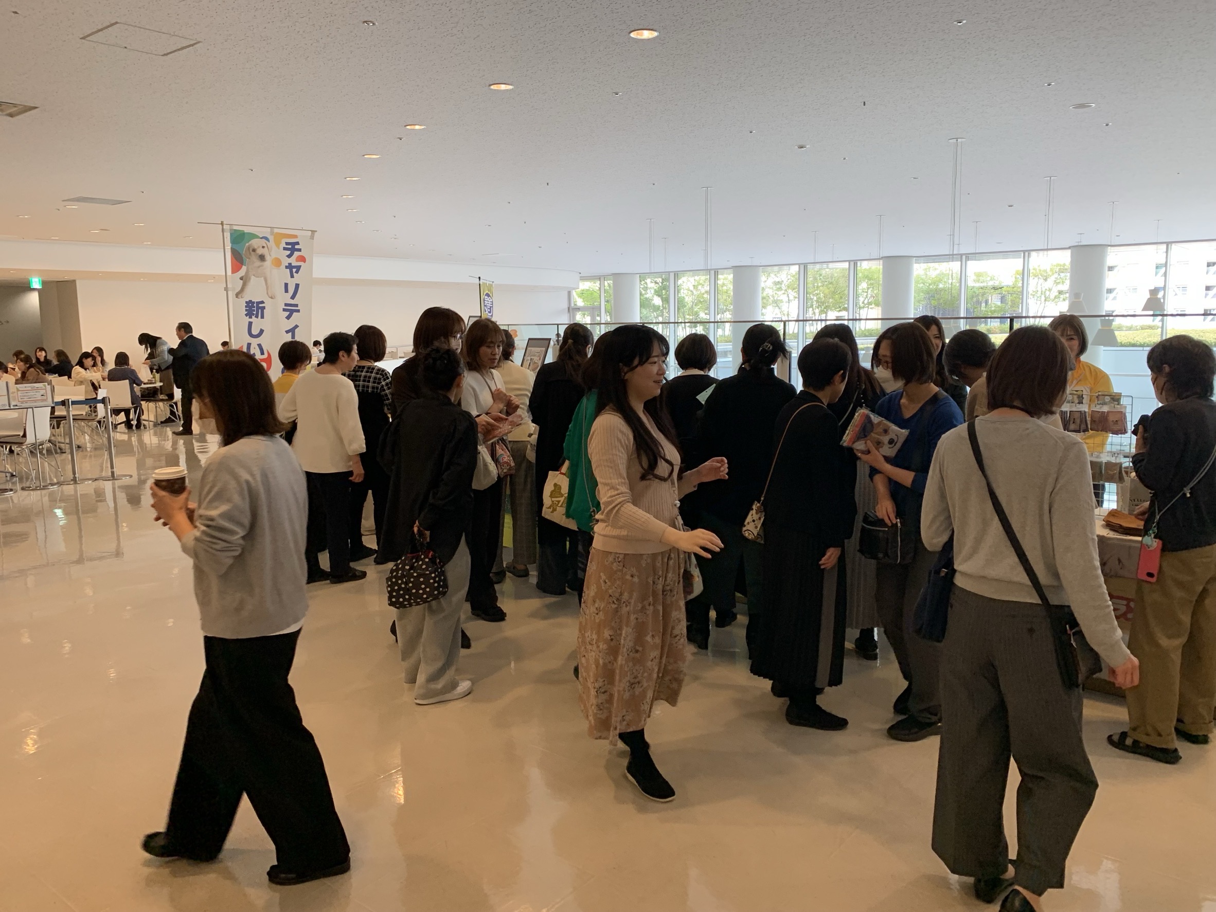 会社の食堂の一角に設置された販売会会場の様子。多くの来場者の後ろ姿が写されている。グッズが並ぶテーブルやスタッフは全く見えない。