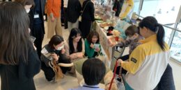 販売会でのふれあいの様子、盲導犬はハーネスを外し、淡いグリーンのニットを着ている。ユーザーが椅子に座り身を乗り出すようにして笑顔。犬の周りを笑顔で数名の来場者が囲んでいる。