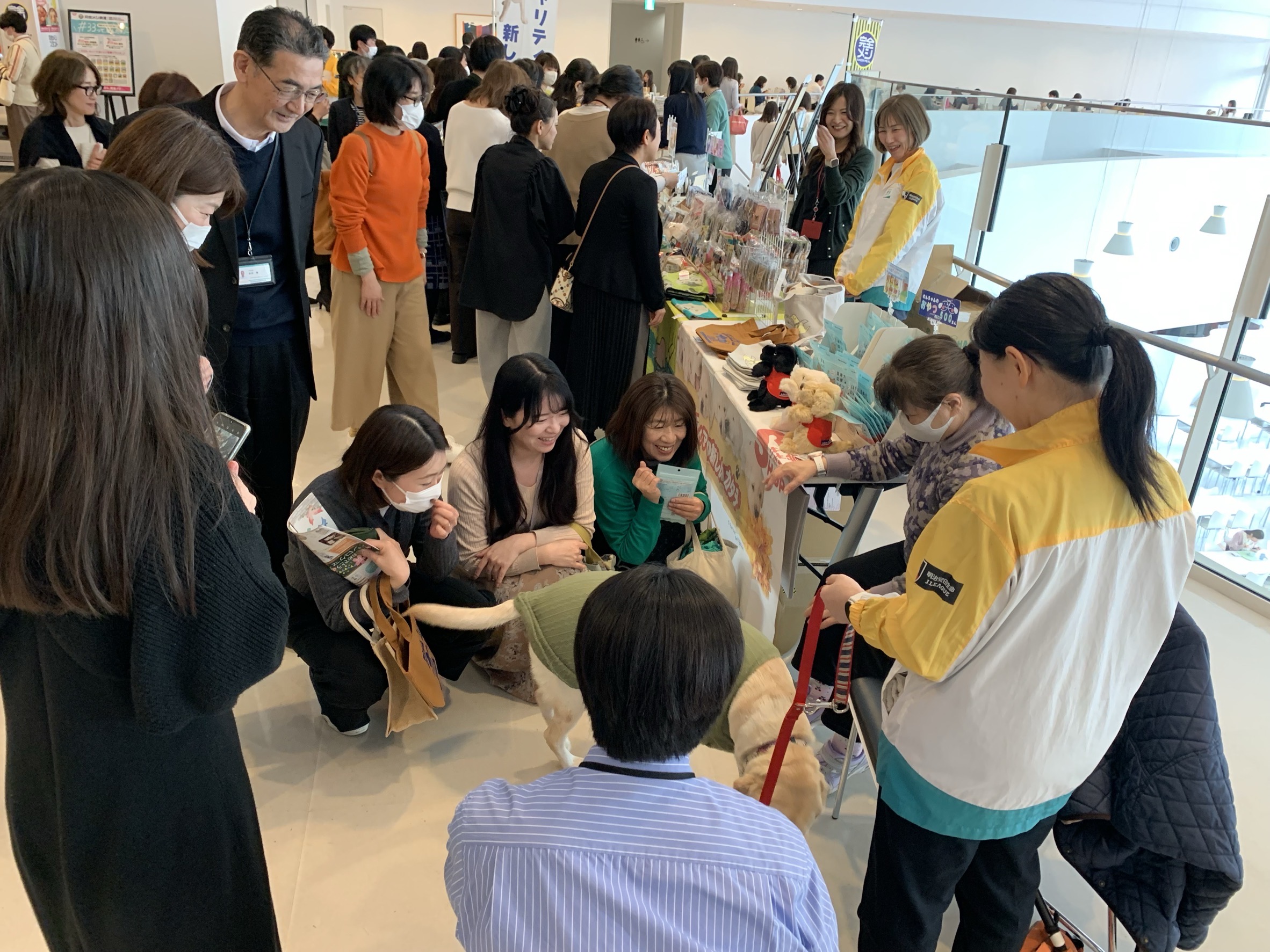 販売会でのふれあいの様子、盲導犬はハーネスを外し、淡いグリーンのニットを着ている。ユーザーが椅子に座り身を乗り出すようにして笑顔。犬の周りを笑顔で数名の来場者が囲んでいる。