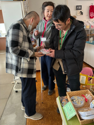 富士ハーネス内のチャリティーグッズショップにて盲導犬ユーザーが買い物をしている。隣で、ファンクラブ参加者がクッキー缶を手渡し説明をしている。