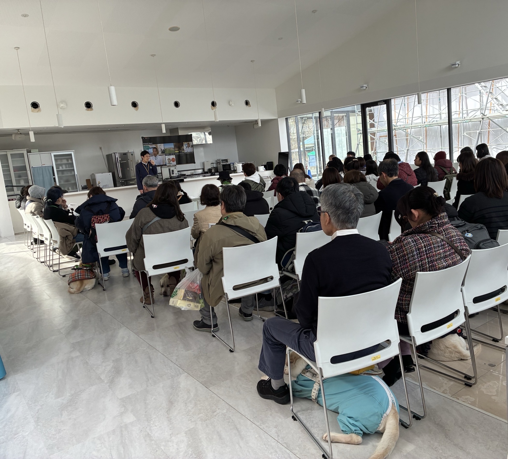 富士ハーネス内でのデモンストレー皆ションの様子。並べられた白い椅子に座り、前方で盲導犬の仕事について説明する協会職員へ顔を向けている。