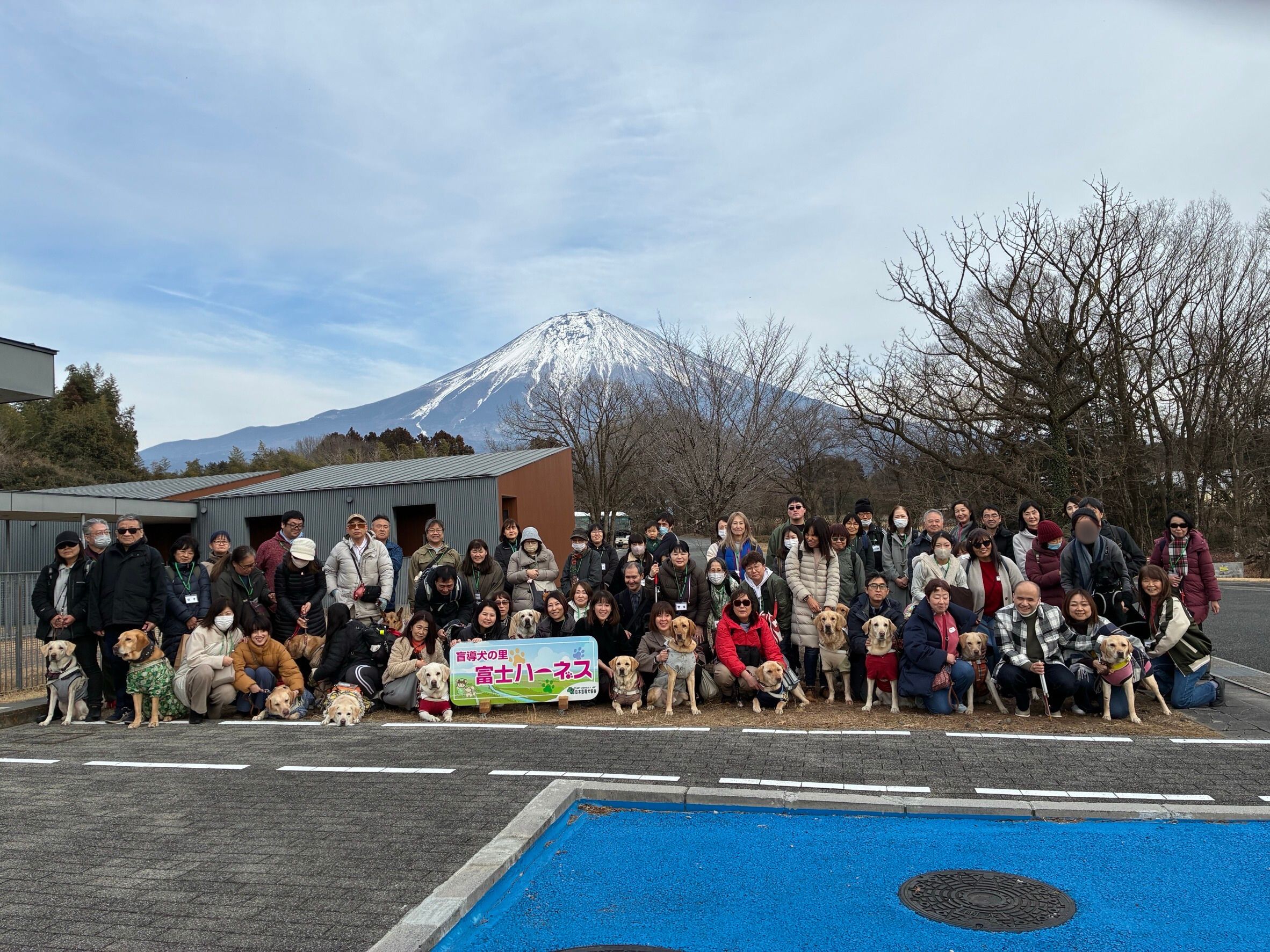 富士ハーネスの外にて。背景には、上部に雪が被った富士山がきれいに見えている。 遠くに見える富士山の裾野の下に、参加者が並んでいる。盲導犬ユーザーと盲導犬が前にしゃがみ、後ろにファンクラブ会員。