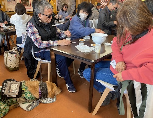 男性の盲導犬ユーザーとファンクラブ参加者が4人掛けのテーブルに座っている。ボトルの中に入った、完成したバターを小さなスプーンですくい、クラッカーにつけ味見をしている。ユーザーの足元には、眠っている盲導犬の頭が見える。