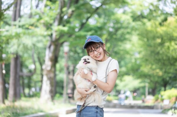緑道を背景に、ポメラニアンのきなこちゃんを抱き、笑顔のけいさん。ブルーのキャップを被り、眼鏡をかけている。